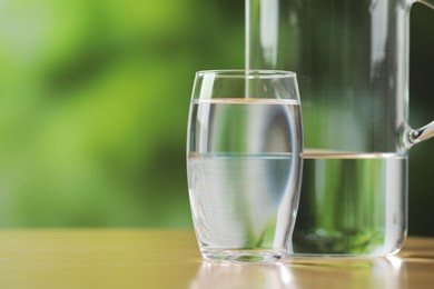 Jug and glass with clear water on table against blurred green background, closeup. Space for text