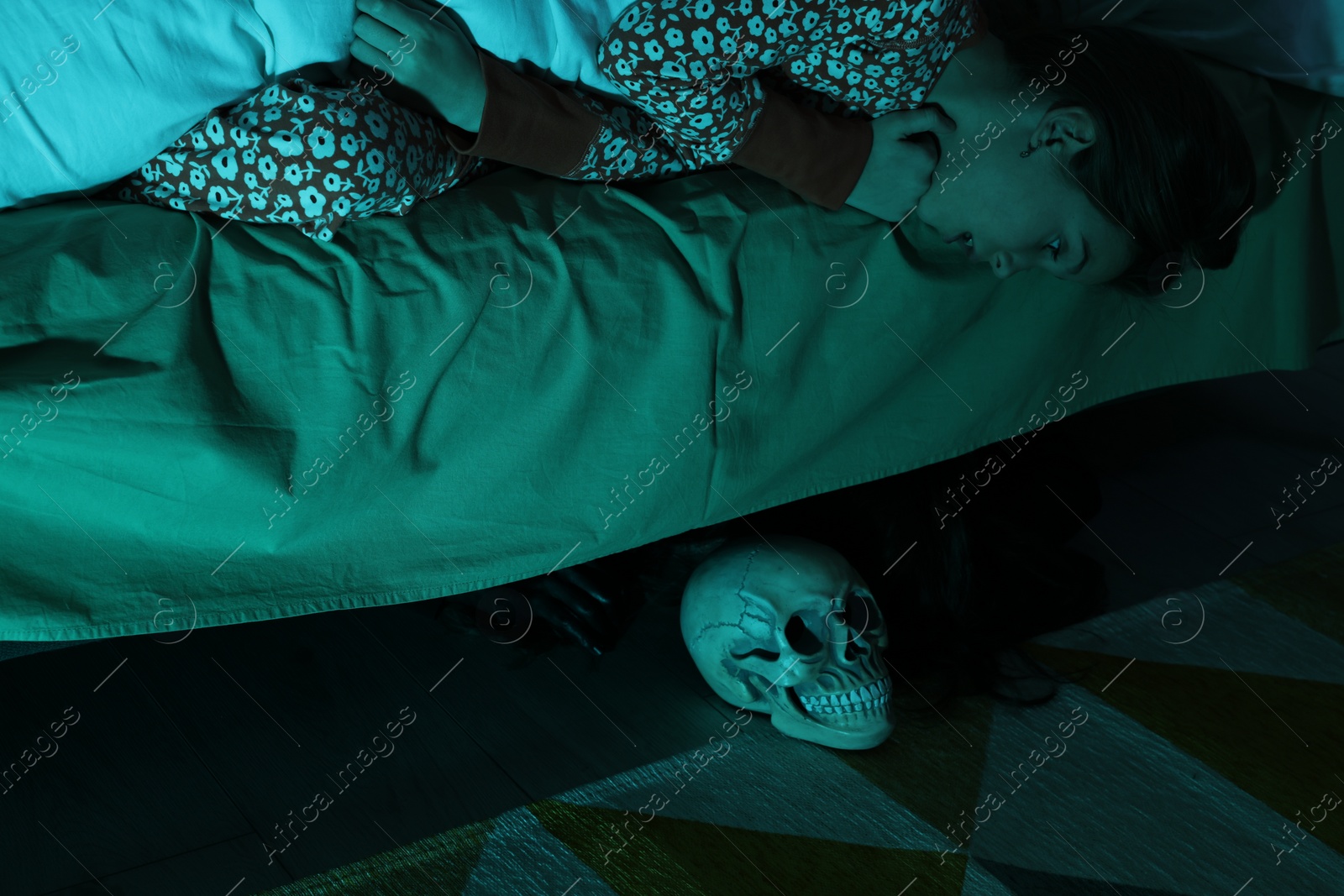 Photo of Childhood phobia. Girl looking at skull under bed indoors