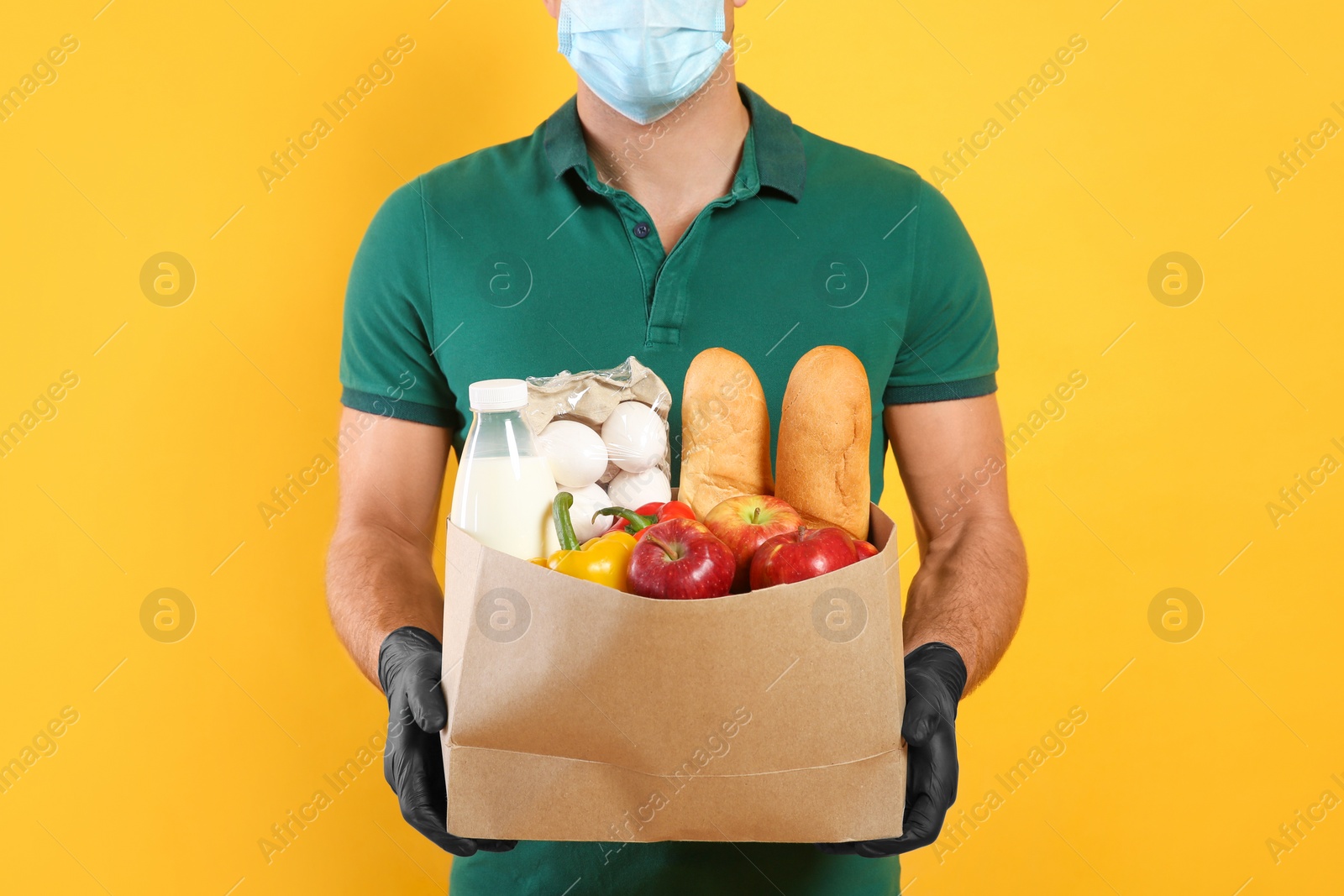 Photo of Courier in medical mask holding paper bag with food on yellow background, closeup. Delivery service during quarantine due to Covid-19 outbreak