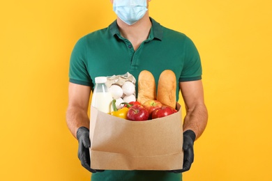 Courier in medical mask holding paper bag with food on yellow background, closeup. Delivery service during quarantine due to Covid-19 outbreak