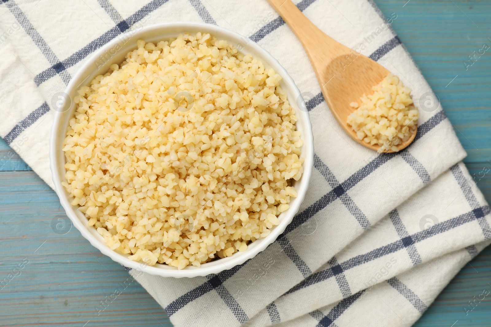 Photo of Delicious bulgur in bowl and spoon on light blue wooden table, top view