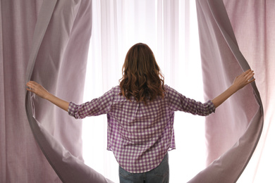 Woman opening window curtains at home in morning, back view
