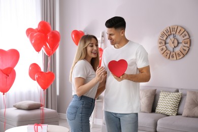 Man presenting gift to his girlfriend in room decorated with heart shaped balloons. Valentine's day celebration