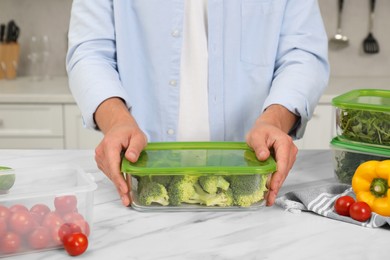 Man holding glass container with fresh broccoli at white marble table in kitchen, closeup. Food storage