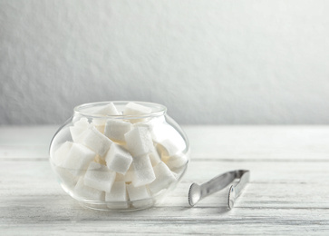 Photo of Refined sugar cubes in glass bowl and tongs on white wooden table. Space for text