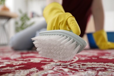 Photo of Woman in rubber gloves cleaning carpet with brush indoors, closeup