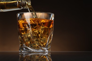 Photo of Pouring whiskey from bottle into glass with ice cubes at table against brown background, space for text