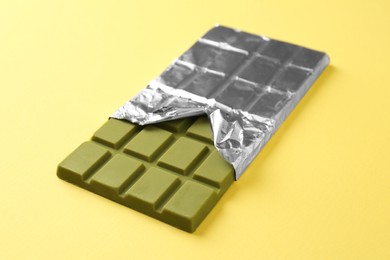 Photo of Tasty matcha chocolate bar wrapped in foil on yellow background, closeup