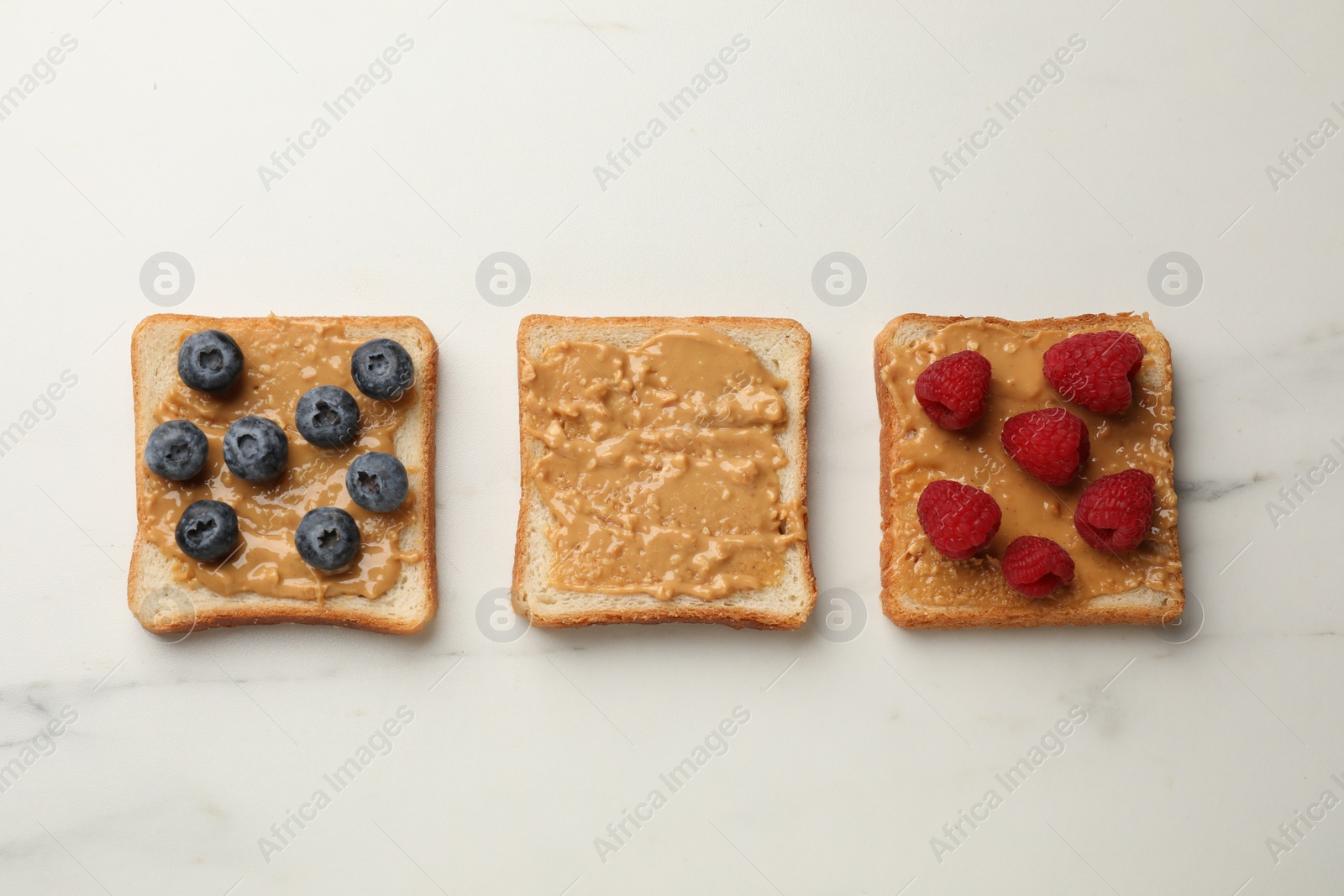 Photo of Delicious toasts with peanut butter, raspberries and blueberries on white marble table, flat lay