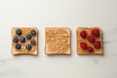 Photo of Delicious toasts with peanut butter, raspberries and blueberries on white marble table, flat lay