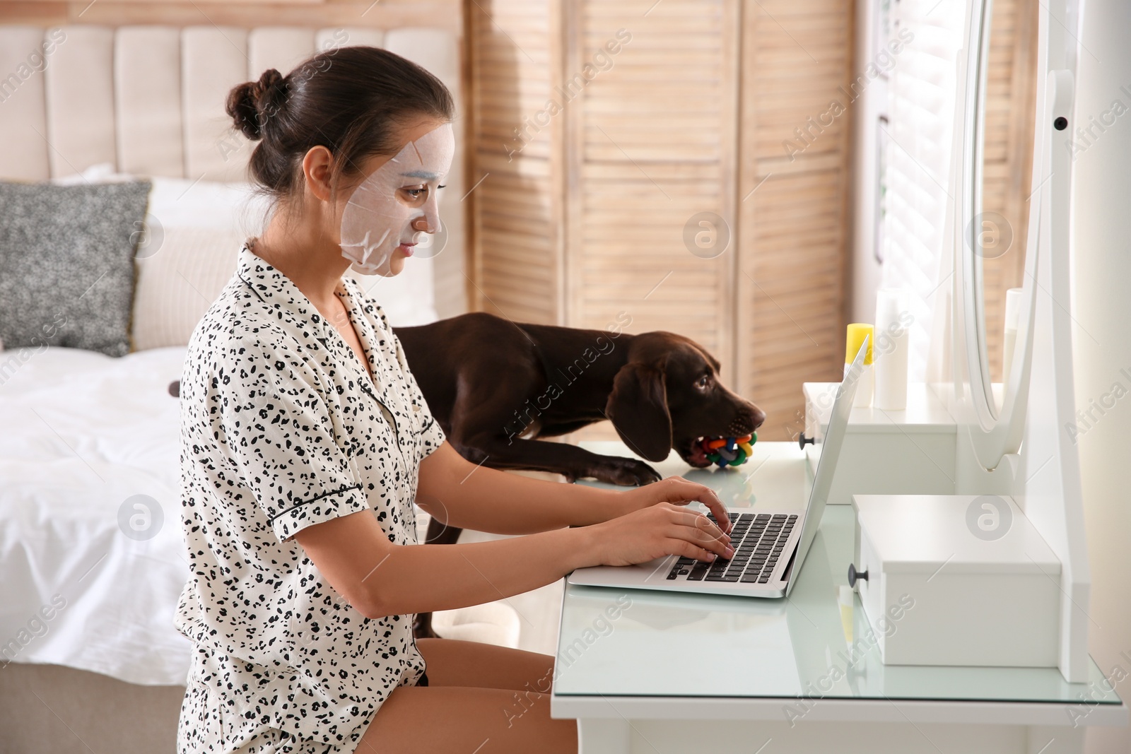 Photo of Young woman with facial mask working on laptop near her playful dog in home office