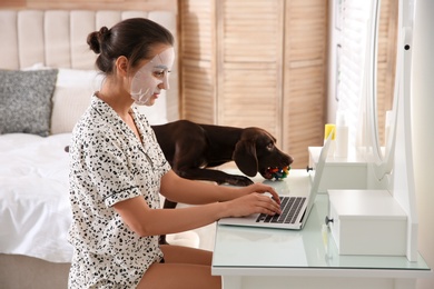 Young woman with facial mask working on laptop near her playful dog in home office