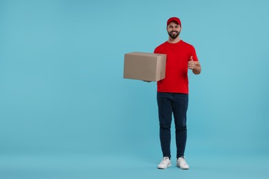 Photo of Happy young courier with parcel showing thumb up on light blue background, space for text