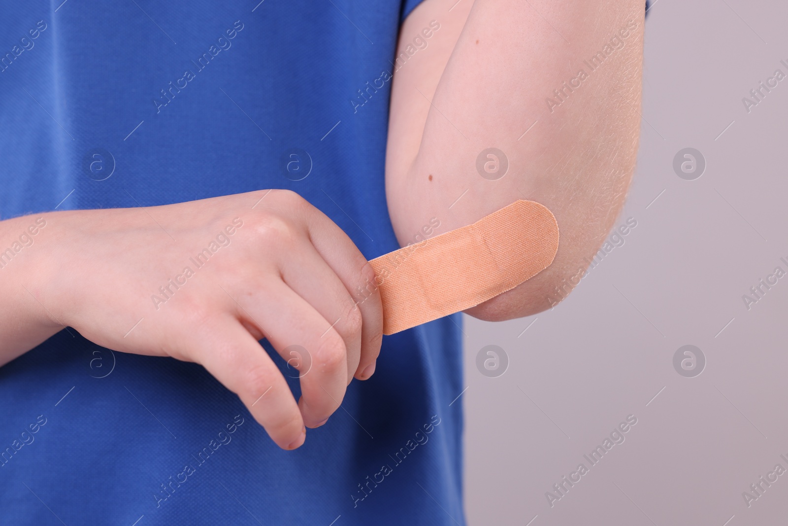 Photo of Little boy putting sticking plaster onto elbow against light grey background, closeup