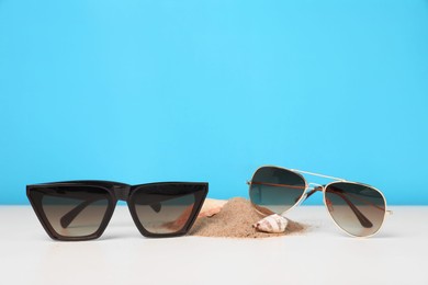 Photo of Stylish sunglasses, seashells and sand on white table against light blue background. Space for text