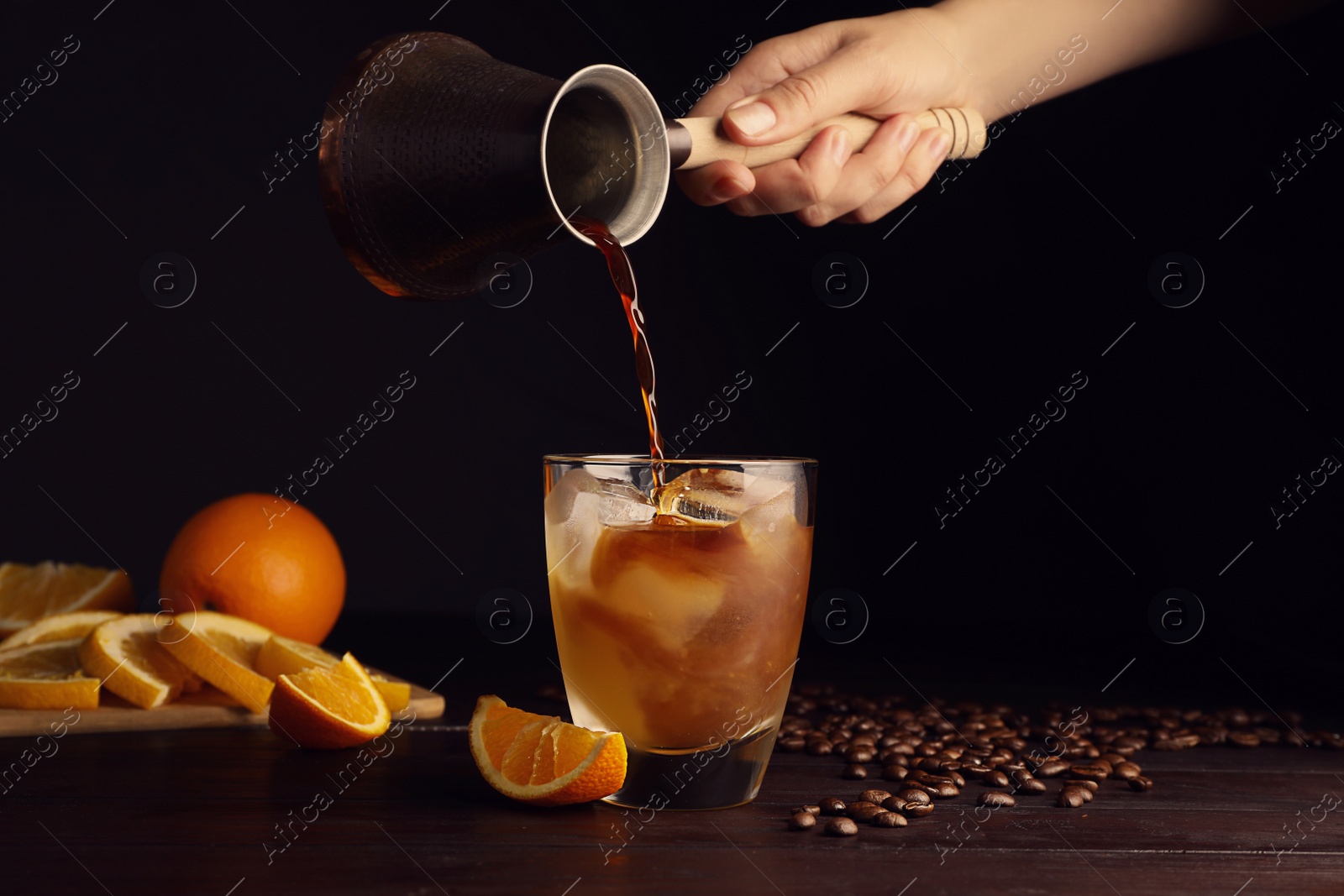 Photo of Woman pouring coffee into glass with orange juice and ice cubes at wooden table, closeup