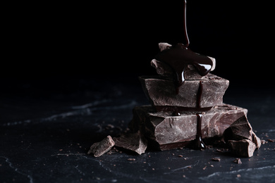 Photo of Pouring syrup onto pieces of dark chocolate on black table, closeup
