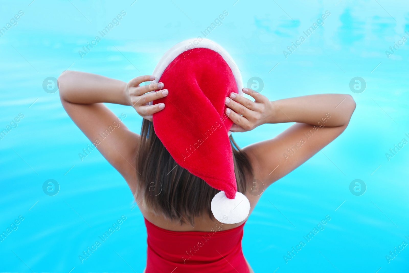 Photo of Young woman wearing Santa Claus hat near swimming pool, back view. Christmas vacation