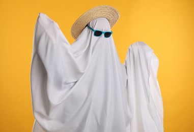 Person in ghost costume, sunglasses and straw hat on yellow background