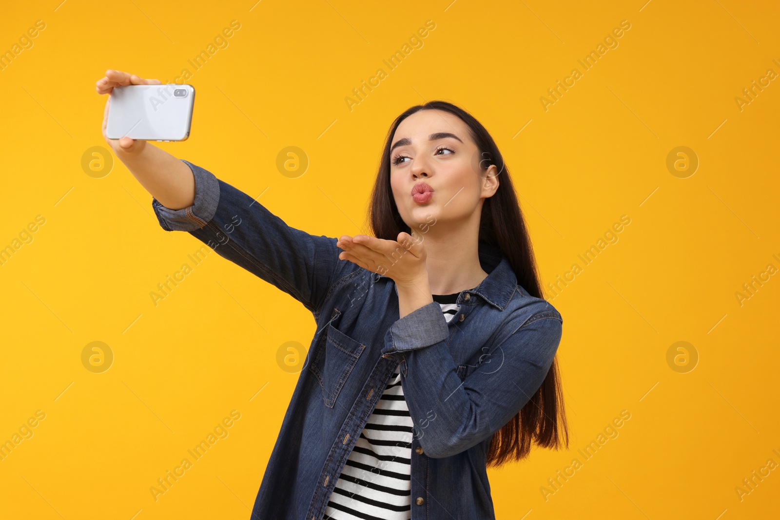 Photo of Young woman taking selfie with smartphone and blowing kiss on yellow background