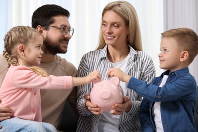 Photo of Family budget. Children putting coins into piggy bank while their parents watching at them indoors
