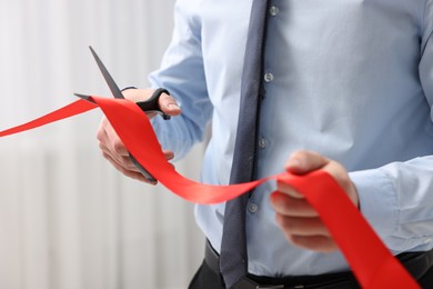 Photo of Man cutting red ribbon with scissors indoors, closeup