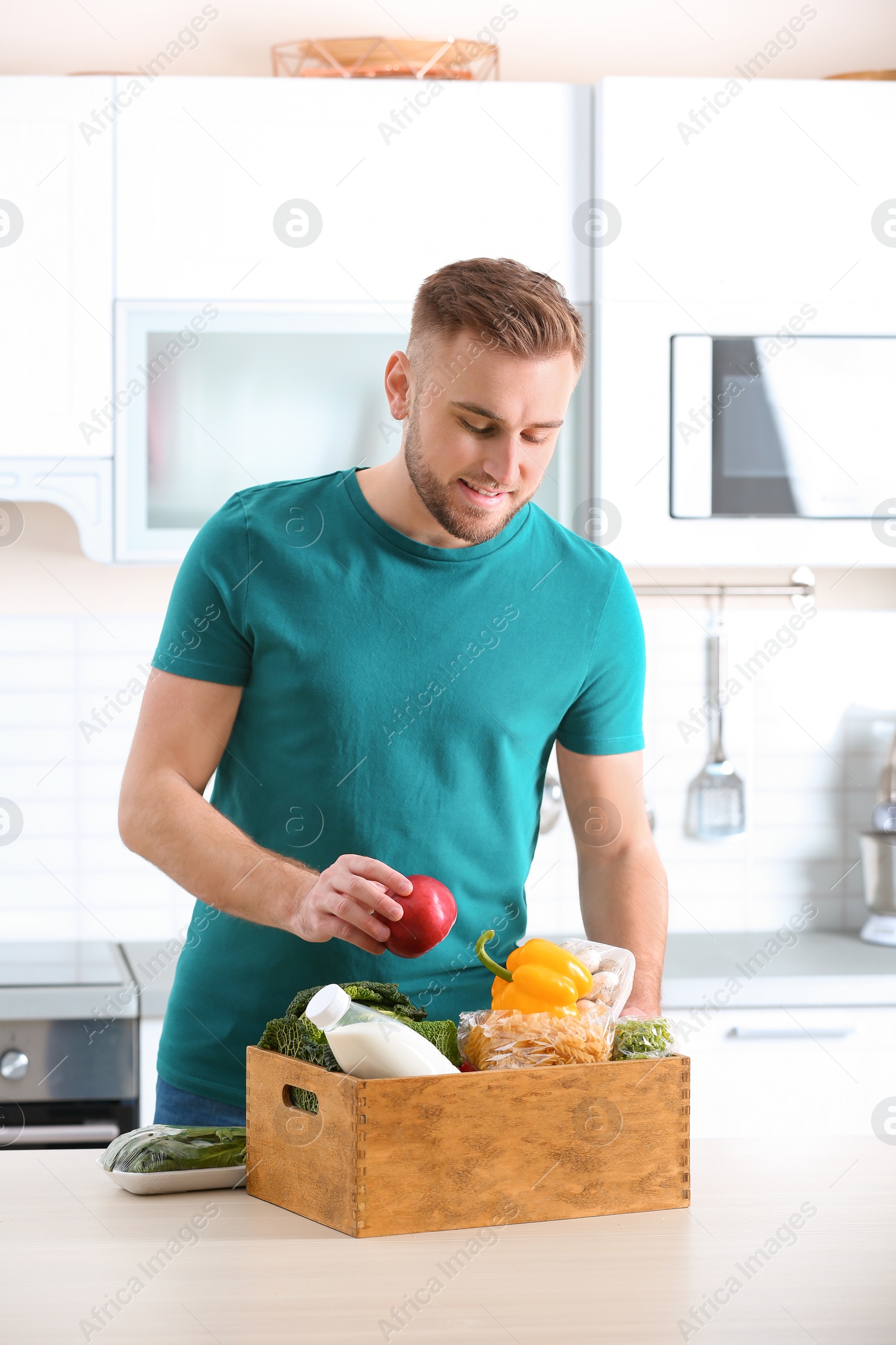 Photo of Young man with wooden crate full of products at table in kitchen. Food delivery service