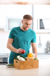 Young man with wooden crate full of products at table in kitchen. Food delivery service