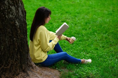 Young woman with cup of coffee reading book near tree in park
