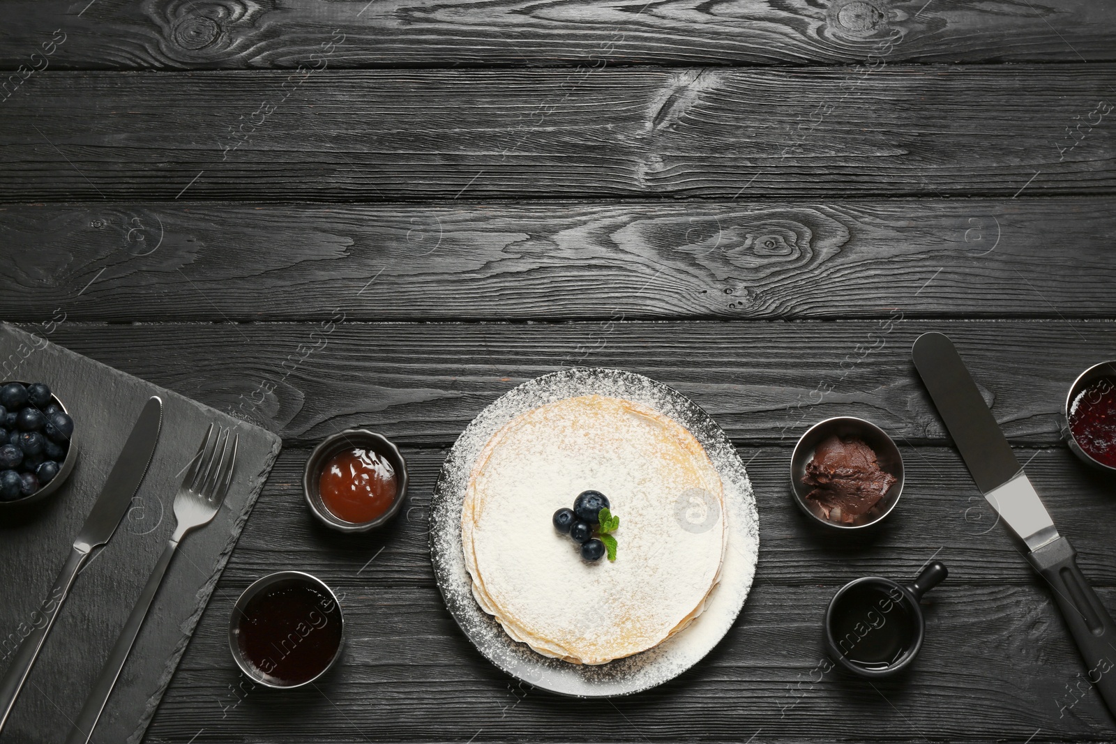 Photo of Delicious crepes with blueberries and different jams on black wooden table, flat lay. Space for text