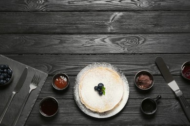 Delicious crepes with blueberries and different jams on black wooden table, flat lay. Space for text