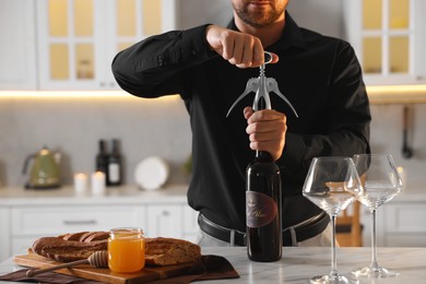 Photo of Romantic dinner. Man opening wine bottle with corkscrew at table in kitchen, closeup