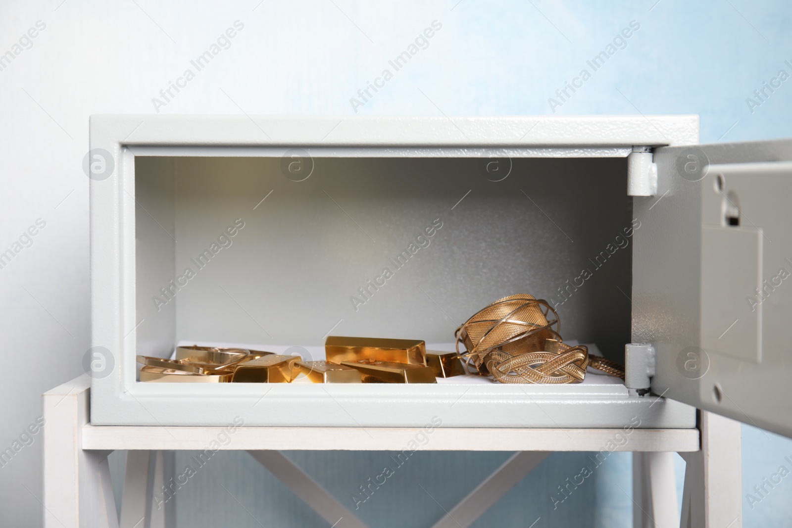Photo of Open steel safe with gold bars and jewelry on white table against light blue background