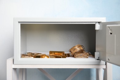 Open steel safe with gold bars and jewelry on white table against light blue background