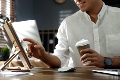 Photo of Freelancer with cup of coffee working on tablet at table indoors, closeup