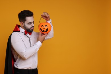 Photo of Man in scary vampire costume with fangs and pumpkin bucket on orange background, space for text. Halloween celebration