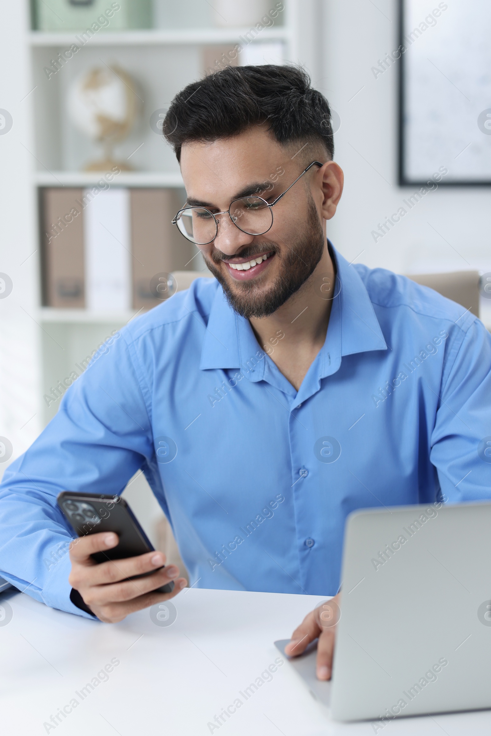 Photo of Happy young man using smartphone while working with laptop at white table in office