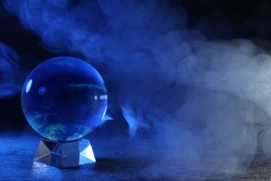 Photo of Magic crystal ball on table and smoke against dark background, space for text. Making predictions
