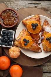 Photo of Fresh tasty puff pastry with sugar powder, jam, tangerines and blueberries on wooden table, flat lay