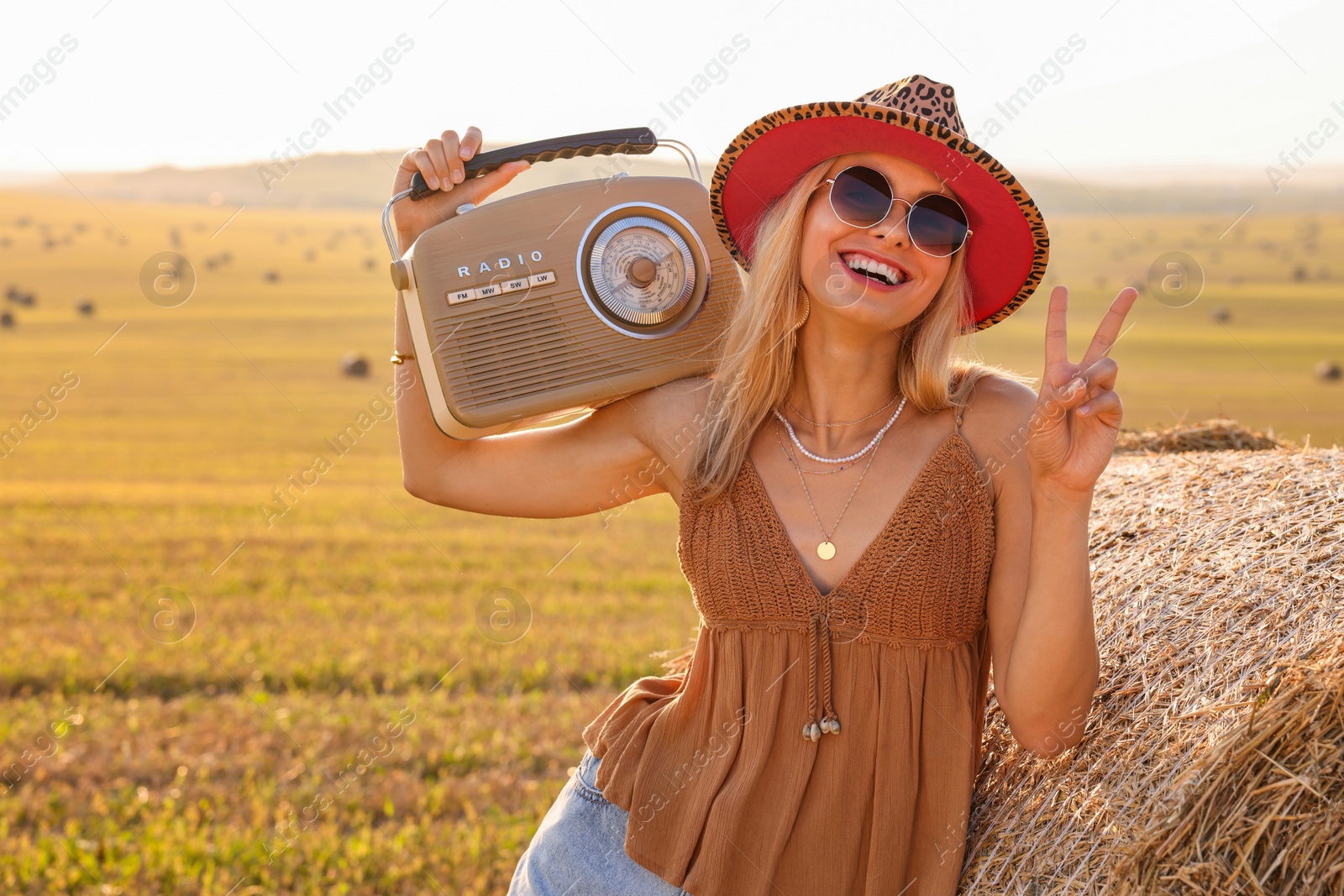 Photo of Happy hippie woman with radio receiver showing peace sign near hay bale in field, space for text