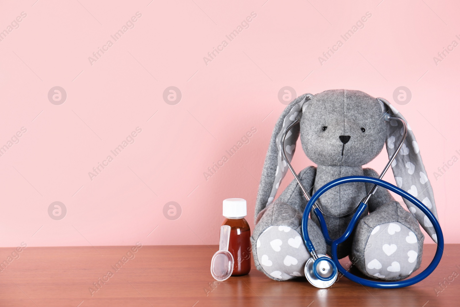 Photo of Toy bunny with stethoscope and cough syrup on table against color background, space for text. Children's hospital