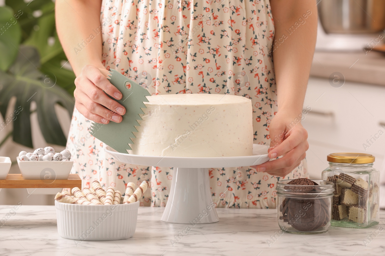 Photo of Woman using scraper to decorate cake at white marble table in kitchen, closeup