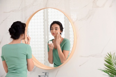 Young woman applying cream on face near mirror in bathroom