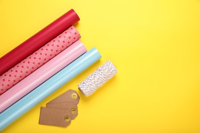 Rolls of colorful wrapping papers, thread and tags on yellow background, flat lay. Space for text