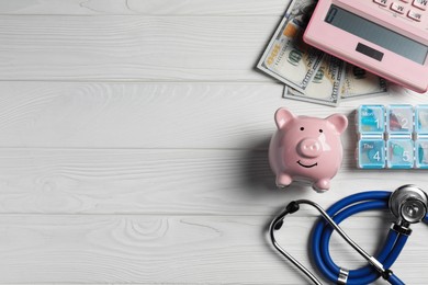 Pale pink ceramic piggy bank, stethoscope and pills on white wooden table, flat lay with space for text. Medical insurance