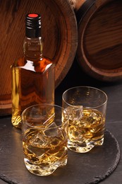 Whiskey with ice cubes in glasses, bottle and wooden barrels on black table