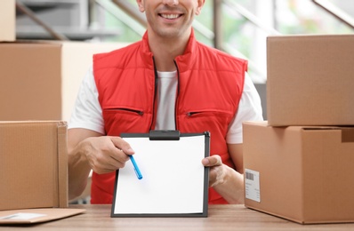 Young courier with clipboard among parcels at delivery department