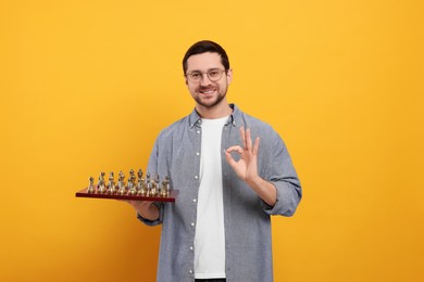 Photo of Smiling man holding chessboard with game pieces and showing OK gesture on orange background