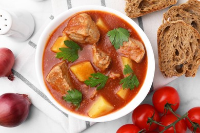 Photo of Delicious goulash in bowl, bread and ingredients on white marble table, flat lay