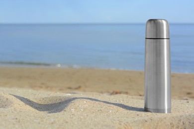 Metallic thermos with hot drink on sandy beach near sea, space for text